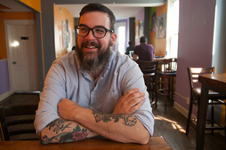 It's him again, but now he's sitting on table with his arms crossed. His sleeves are rolled up and his forearms are tattooed.